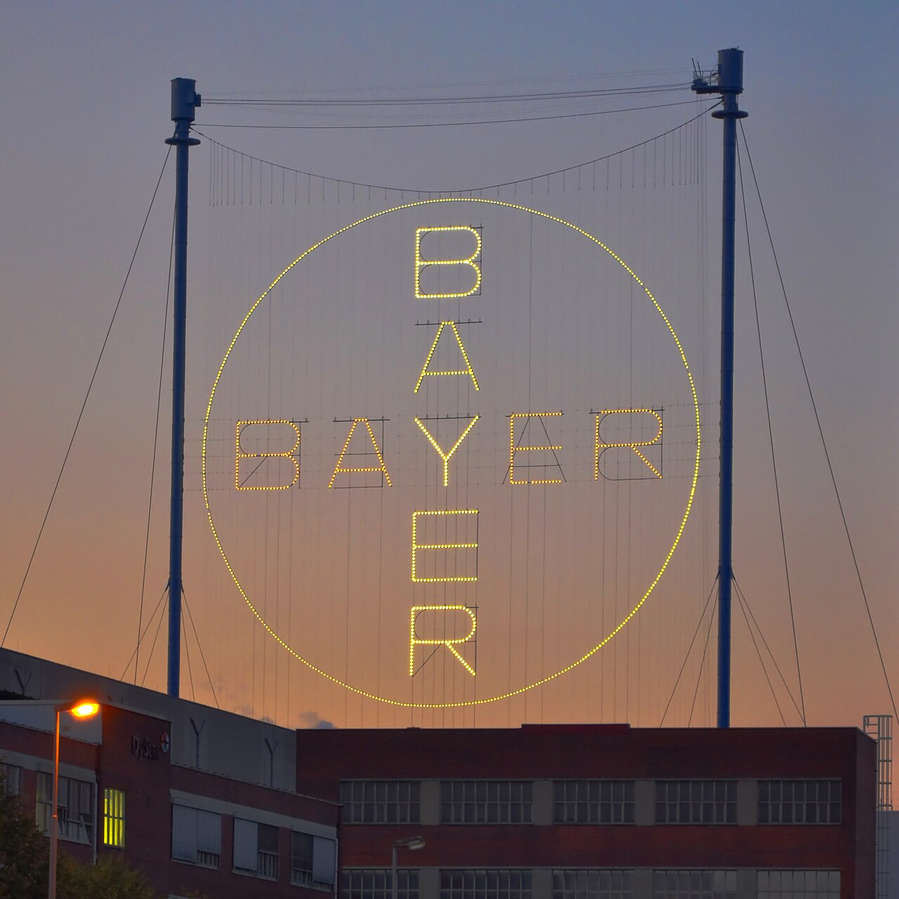 La croix Bayer, symbole de la marque, éclaire la nuit à Leverkusen. Un emblème désormais indissociable du plus grand scandale sanitaire et juridique de l'histoire de l'entreprise.